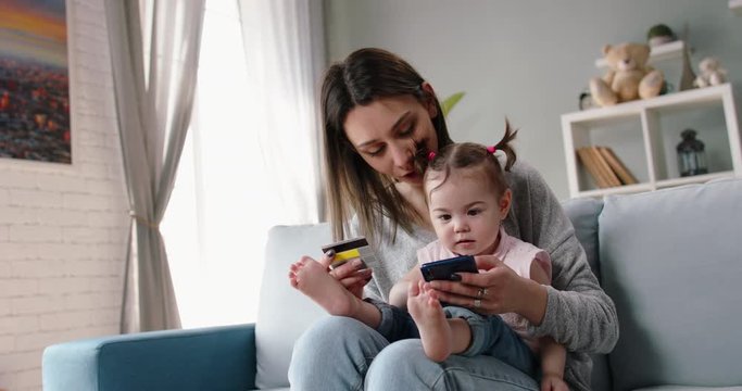 Relaxed caucasian mom is making an online purchase with credit card using smartphone while holding her cute little baby girl on knees - happy family, technology, finance 4k footage