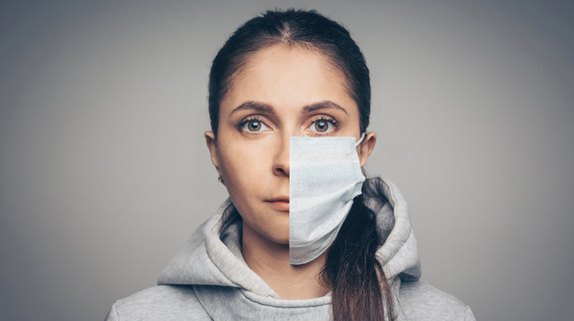 Studio Portrait Of Young Woman Wearing A Face Mask, Looking At Camera, Close Up On Gray Background. One Half Face In Mask Another Without