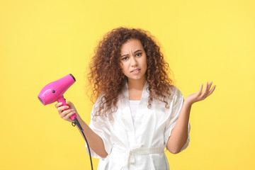 Beautiful confused African-American woman with blow dryer on color background