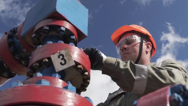 Oilman in orange helmet and protective glass opening valve of x-mass wellhead to shut off well. Cross-type Christmass tree and high pressure piping destined for wellhead sealing. Extraction of oil.