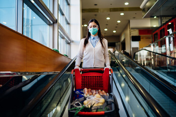 Woman wearing mask groceries shopping in supermarket,pushing trolley.Food panic buying and hoarding.Covid-19 quarantine shopper with shopping cart.Sustainable coronavirus consumer restrictions