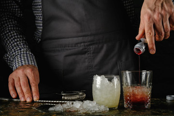 The bartender makes a cocktail, pours red liquid from a bottle into a glass with ice. Near ice and a bar spoon.