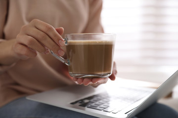 Woman with cup of coffee and laptop indoors, closeup