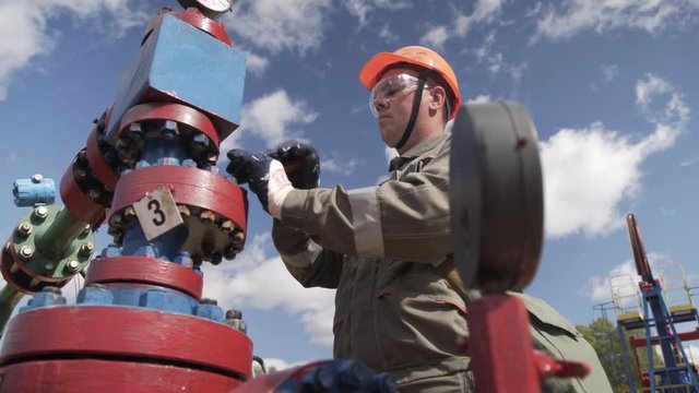 Oil worker in orange helmet and protective glass opening or closing valve of x-mass wellhead to shut off well. Cross-type Christmass tree and high pressure piping destined for wellhead sealing.