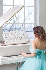 young woman with curly hair in aquamarine dress playing piano in white room