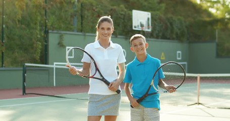 Portrait of Caucasian beautiful mother and her little cute son looking at each other, smiling to camera and standing at tennis pitch. Young pretty female instructor and small boy with rackets in hands