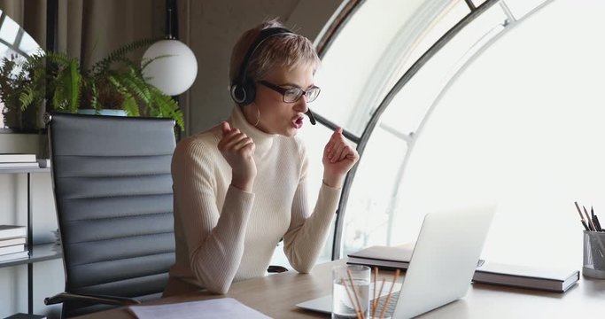 Young business woman wearing headset conferencing in distance remote web chat sitting at office desk. Female worker, coach, manager participating webcam video call speaking looking at laptop.