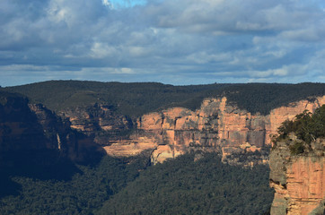 Fototapeta na wymiar Sandstone cliffs are bathed in sunlight. They are surrounded by forest. The sky is overcast. They form part of the Megalong Valley in the Blue Mountains, Australia.