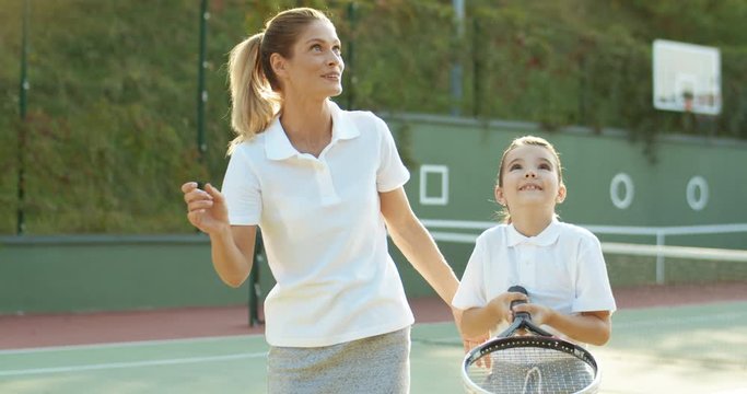 Beautiful sporty young female instructor showing to girl how to play tennis on sport court. Caucasian pretty woman teaching small cute daughter how to hit ball with racket on sunny summer day.