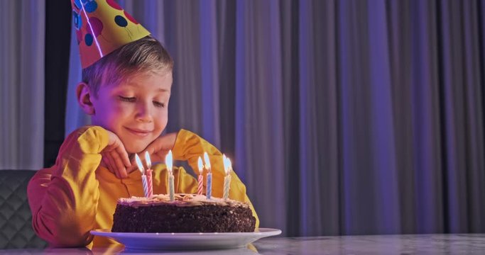 Cute boy blows out candles on a cake on his birthday.  Happy child looks at a cake with candles. Child looks at the cake and makes a birthday wish. Boy celebrates his birthday. Happiness concept