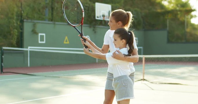 Beautiful sporty young female instructor showing to girl how to play tennis on sport court. Caucasian pretty mother teaching small cute daughter how to hit ball with racket on sunny summer day.