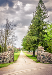 Aged effect - An open gate with stone wall & a long road with soft focus background of a farm and  cloudy sky.