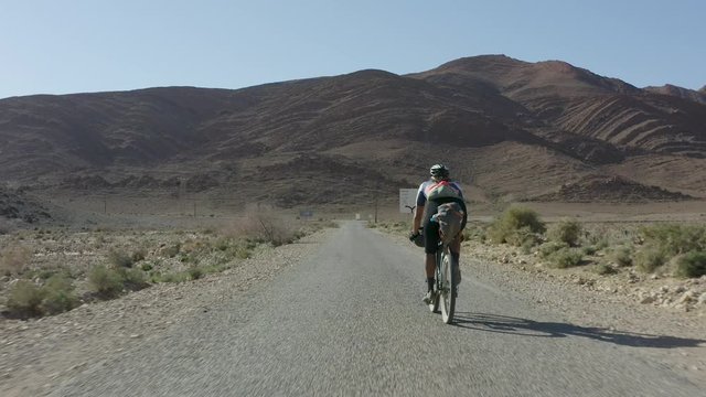 A low aerial POV shot of a female cyclist bikepacking on a paved road through an arid North African desert landscape, passing road signs with Arabic writing on them.