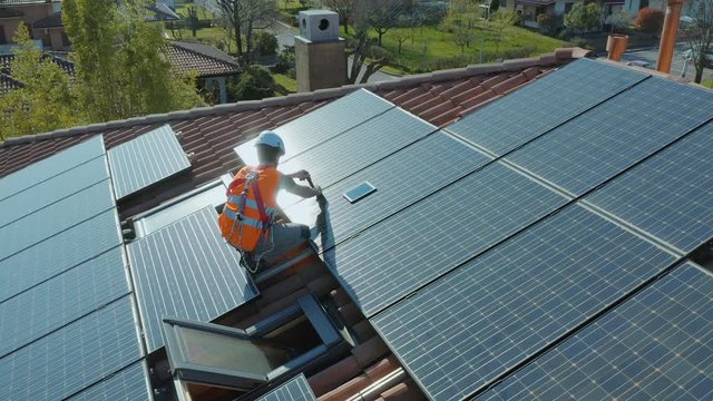 Aerial shot of technician with hard hat and safety equipment installing and working on maintenance of photovoltaic panel system installed on home domestic roof top, urban landscape. Wide angle camera.