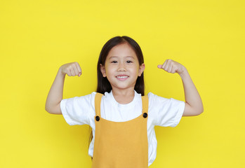 Smiling asian little girl showing his muscle with looking camera isolated on yellow background.