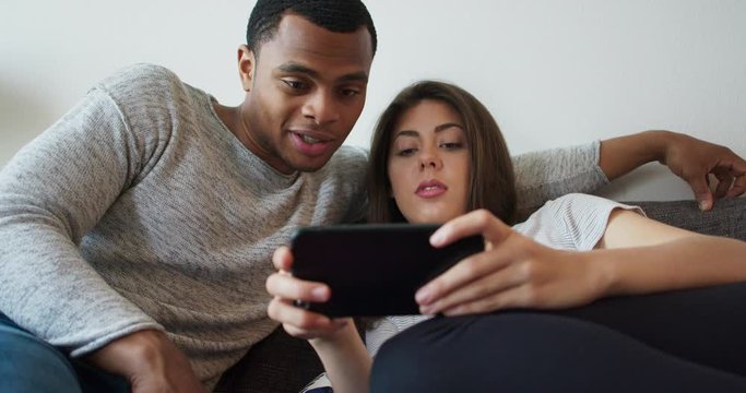 Young millennial couple laughing at a cute video on their phone. African American and Caucasian boyfriend and girlfriend browsing the internet using social media. 4k Slow motion handheld