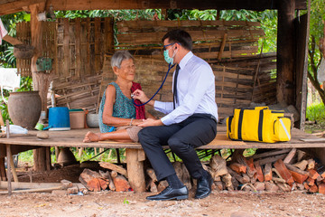 Doctor wearing protective mask to Protect Against Covid-19 listening heart beat and breathing of Elderly Woman with Stethoscope with First Aid Medical Box at Remote Areas. © visoot