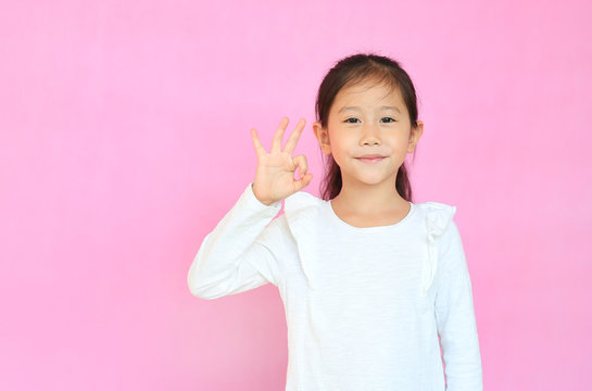 Beautiful Asian Little Child Girl Making Ok Sign Isolated Over Pink Background. Portrait Of Smiling Kid Show Confident Hands Symbol.