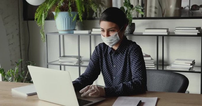 Female indian remote worker wears medical gloves and face mask using laptop computer working from home office on quarantine. Keeping safety recommendations for coronavirus covid 19 protection concept.