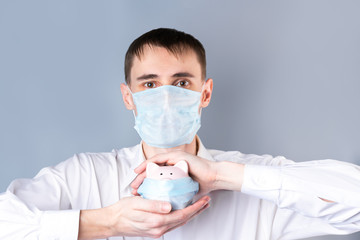 A man in a medical mask holds a pink piggy Bank protected by a medical mask  on a gray background in his hands,  looks at the camera . Coronavirus