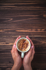 Female hands holding a cup of coffee with foam over a wooden table, top view. Latte or cappuccino with chocolate sprinkles. copy space