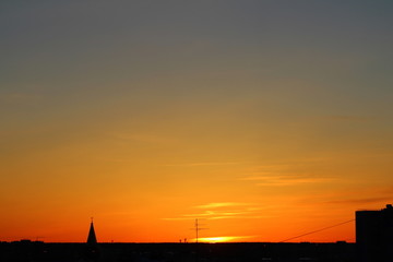 Naklejka premium Sun below the horizon and the roof of the church with a cross, television antenna, roof of a multi-storey urban building in the background in the background fiery dramatic orange sky at sunset or dawn
