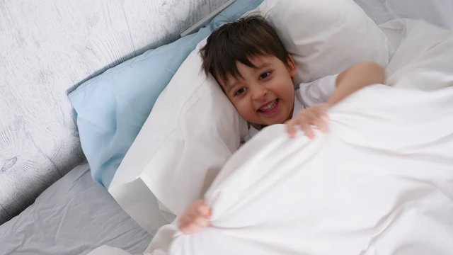 child in a white bed hides under a blanket at home