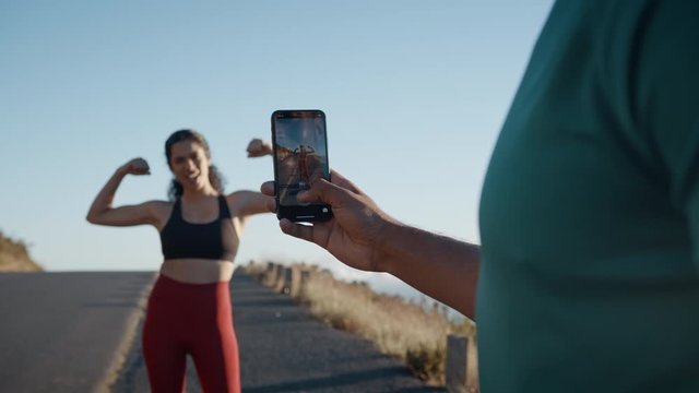 Man taking picture of a woman flexing her biceps on a hillside road in the morning. Man and woman runner taking break after running workout on mountain road taking photos with a mobile phone.
