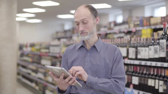 Portrait of professional senior trader using tablet in alcohol supermarket. Confident Caucasian bearded man swiping screen and smiling as standing between rows of beverages in the shop.