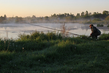 Obraz na płótnie Canvas On a pond covered with a misty haze, at sunrise, a fisherman cast a rod and sat down.