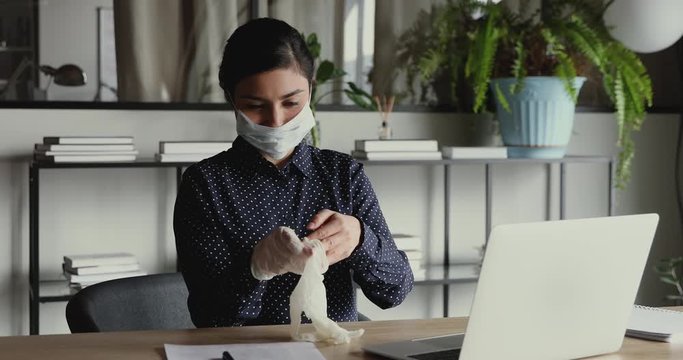 Young indian business woman wearing medical gloves and face mask before using computer working from home office on quarantine. Coronavirus infection spreading prevention, covid 19 protection concept.