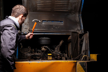 Fototapeta na wymiar An angry male mechanic is repairing the engine of an old truck with an open hood hitting it with a hammer.