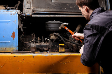 An angry male mechanic is repairing the engine of an old truck with an open hood hitting it with a...