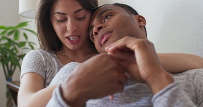 African American and Caucasian millennial couple holding each other on the couch as they talk. Man and woman sharing a lazy afternoon together idly holding hands and chatting. 4k slow motion handheld