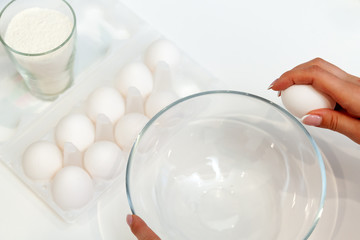 Close-up on the hands of a woman who cracks an egg with her fingers and pours the yolk into a plate against the background of a package with a dozen eggs and a glass with flour