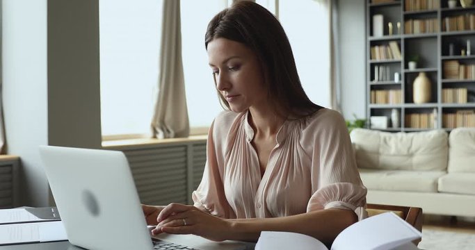 Focused young woman sitting at table, typing message on computer, studying on online courses. Concentrated businesswoman communicating with client or working remotely on laptop from home alone.