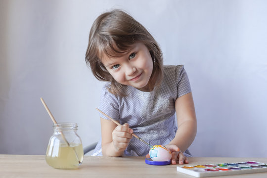 A small smiling girl is sitting at home at the table and paints a white chicken egg with watercolors. She is dressed in a gray dress. On a light gray background. Selective focus