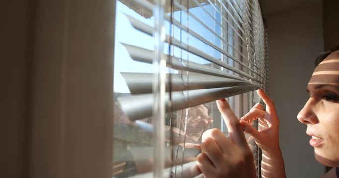 Close up profile portrait from indoors of attractive brunette woman pushing blinds at window with hands. Looking fearfully outside because afraid of germs. The concept of quarantine,  self-isolation