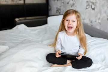 A little girl with blond hair on the bed in the room. Holding a tablet computer, crying. The blonde girl is upset.