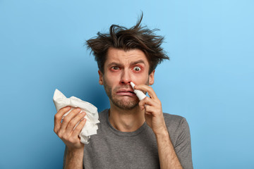 Photo of unhappy European man with runny nose, holds nasal drops and handkerchief, suffers from...