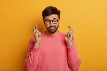 Hopeful bearded man crosses fingers wants get job, searches career opportunities awaits for result after interview stands in anticipation puts all efforts in wishful thinking pleads dream comes true