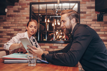 Smiling business partners, young man and woman sitting in cafe, talking positively and using a digital tablet