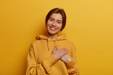 Pretty adult woman feels touched, feels thankful for getting compliment, tilts head and smiles pleasantly, wears casual hoodie, isolated over yellow background, appreciates friends help or advice