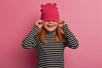 Funny child hides face with pink hat, smiles positively and has playful mood, dressed in striped jumper, enjoys carefree childhood, isolated on pink background, smiles broadly. Naughty daughter