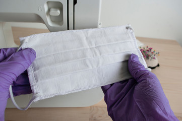 Hands in violet gloves holding mask. Sew machine behind.Protect yourself against coronavirus, sewing medical face mask at home.