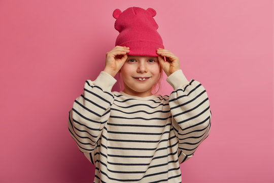 Adorable small child dresses loose striped jumper and pink hat, has two teeth stick out, going to play with friends at playground, expresses positive mood, isolated on pink wall. Childhood concept