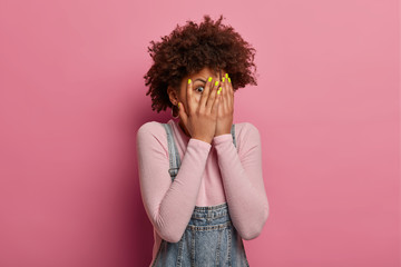 Scared curly woman peeks through fingers, hides face with palms, afraids of terrifying scene as watches horror movie, sees phobia, stands alone, feels frightened, isolated on pink background