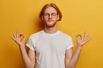 Patient red haired millennial man stays calm, feels relaxed, enjoys peaceful atmosphere, keeps eyes closed, stands in lotus pose, makes zen gesture, wears white t shirt, poses against yellow wall