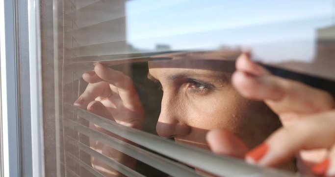 Close up front portrait from outdoors of attractive brunette woman pushing blinds at window with hands. Looking fearfully outside because afraid of germs. The concept of quarantine,  self-isolation