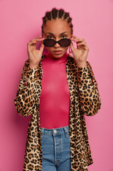 Vertical shot of serious looking fashion model looks through sunglasses, wears leopard shirt and jeans, poses for magazine cover, isolated on pink background. People, style and beauty concept